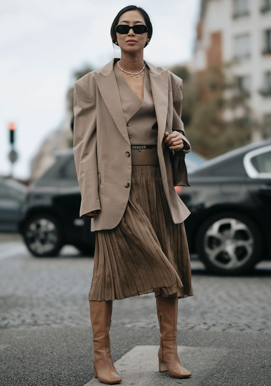 Frau trägt ein monochromes Outfit bestehend aus beigem Plisseerock und kombiniert dazu einen beigen Blazer